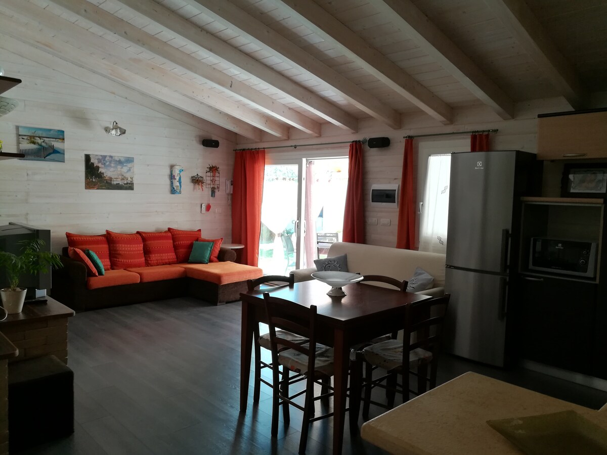 A spacious living area features a comfortable seating arrangement with bright orange cushions. A wooden dining table is surrounded by chairs, and large windows invite natural light into the room. The kitchen area, equipped with modern appliances, is easily accessible from this space.