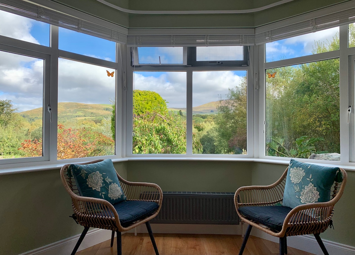 A sunlit bay window area features two wicker chairs adorned with decorative cushions. Panoramic views of the lush green countryside and rolling hills can be seen through the large windows, inviting the outdoors inside.