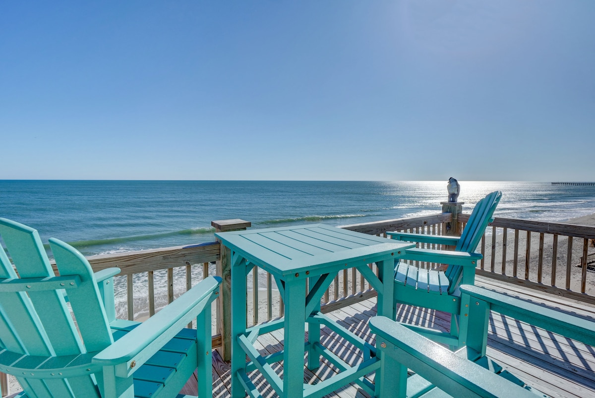 Four turquoise chairs surround a matching table on a wooden deck, overlooking a serene ocean view. The calm water reflects sunlight, with soft waves gently lapping at the shore in the background. A clear blue sky enhances the peaceful coastal atmosphere.