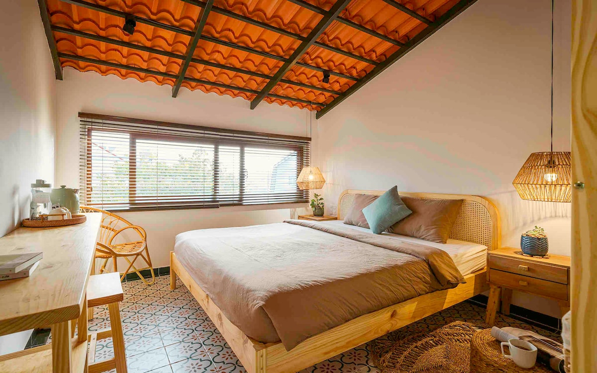 A cozy bedroom features a spacious bed with soft, neutral bedding, complemented by decorative pillows. Natural light fills the room through large windows with wooden blinds. A wooden desk and chair, along with woven lamps, provide a relaxed aesthetic, while patterned flooring adds character.