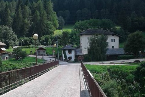 A stone bridge is shown spanning a green valley, with trees lining the horizon. A few buildings are visible in the background, surrounded by lush vegetation. Street lamps line the bridge, providing a gentle illumination to the pathway.