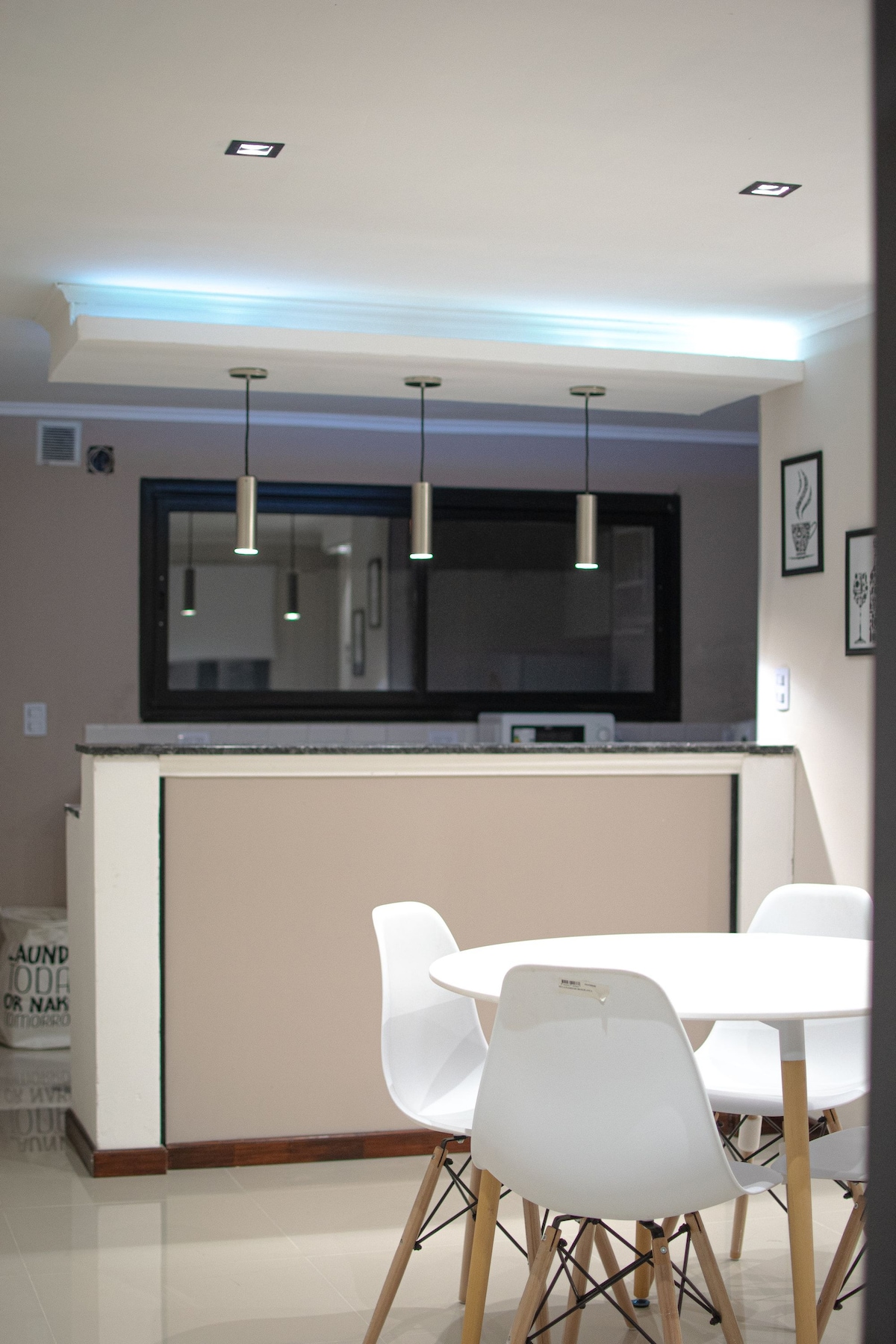 A modern kitchen area is visible with a white circular table surrounded by four sleek chairs. Pendant lights hang above the table, providing focused illumination. A window with a black frame reflects the organized countertop, and a microwave is positioned on the back ledge.