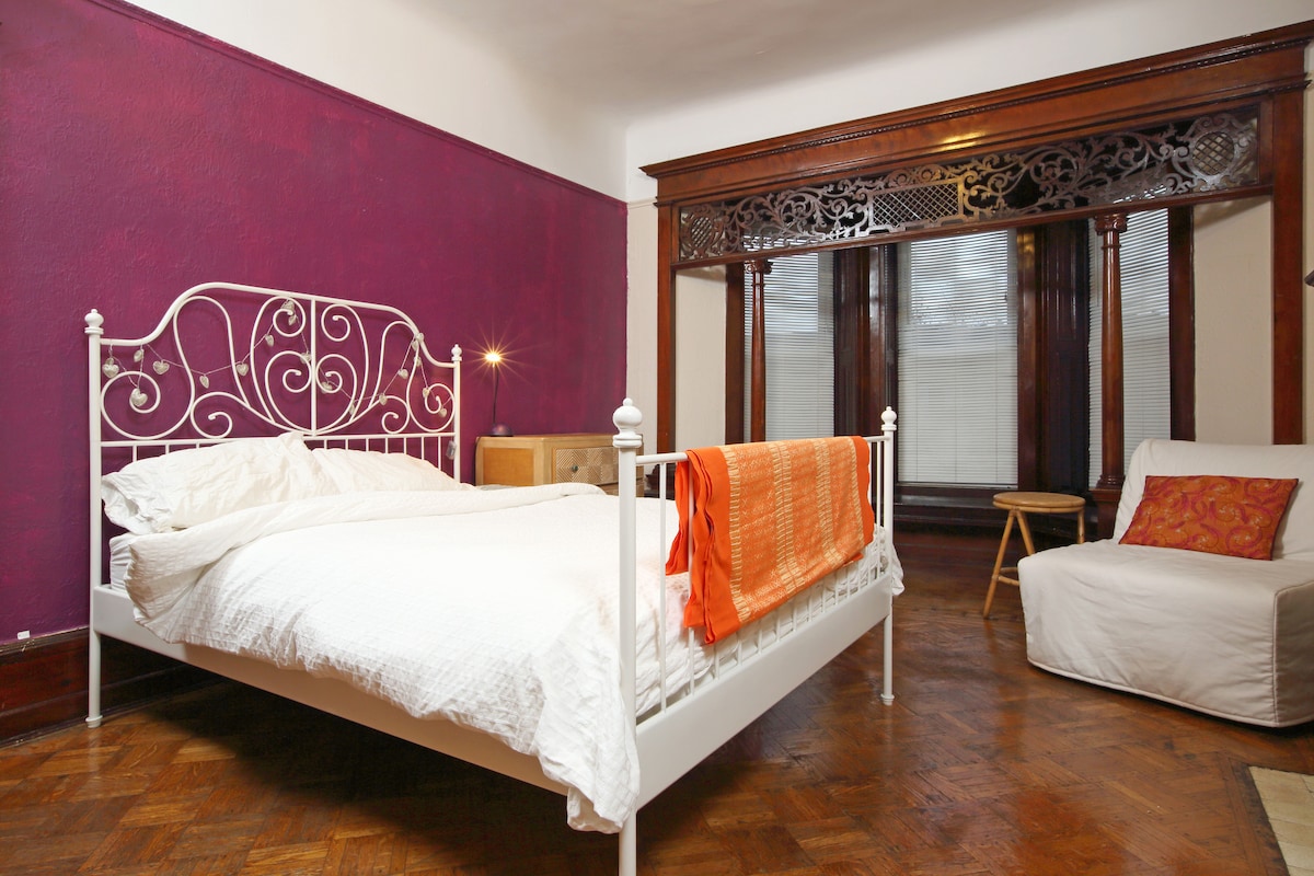 A bedroom features a white iron bed adorned with white bedding and an orange throw. A cozy chair sits in the corner. Dark wood accents highlight the windows, allowing soft natural light to filter through the sheer curtains.