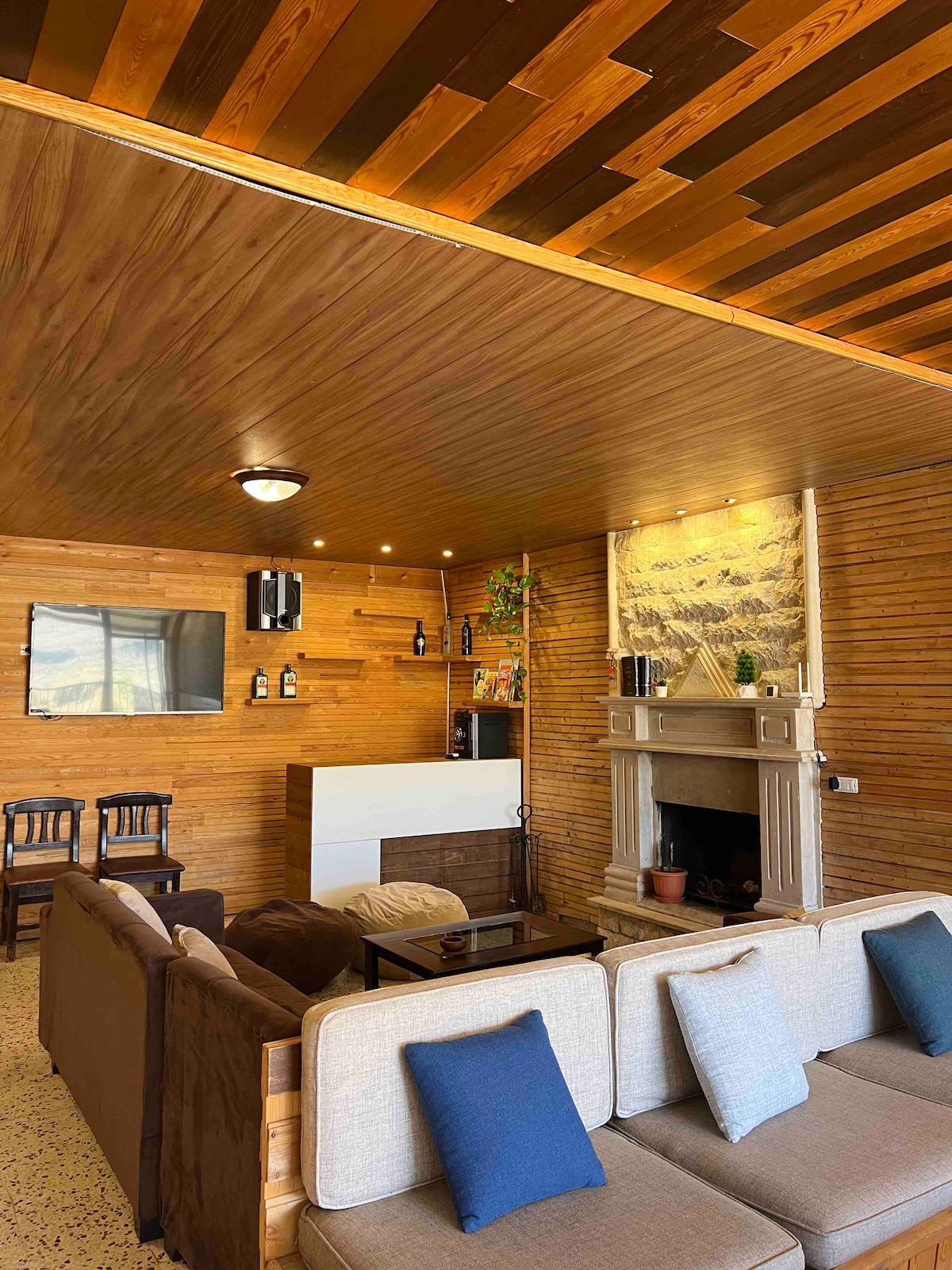 A warm living area is highlighted by wood-paneled walls and a wooden ceiling. A plush sectional sofa is arranged around a coffee table, with decorative cushions providing comfort. A stone fireplace adds a cozy focal point, complemented by a sleek white mantel and shelves adorned with bottles and plants.