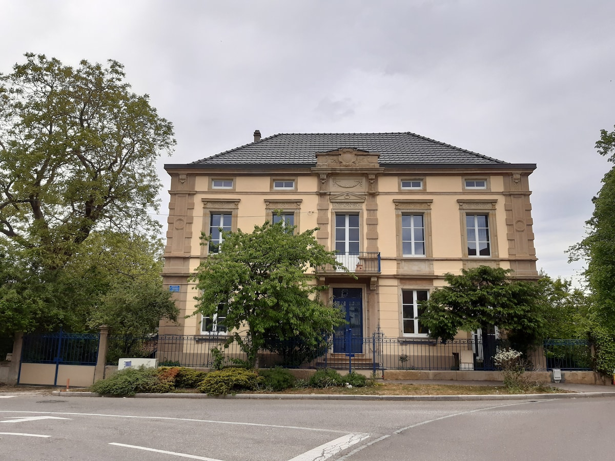 An elegant 19th-century mansion is captured from the street view, showcasing classic architecture with large windows and intricate detailing above the entrance. Green trees and shrubs frame the exterior, while a blue door adds a pop of color to the facade.