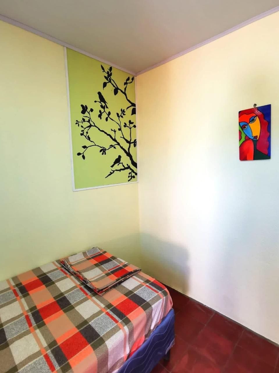 A cozy corner of the room features a spacious bed with a checked blanket. A vibrant green wall is adorned with a simple bird motif, while a colorful artwork hangs nearby, adding a touch of creativity to the serene space.