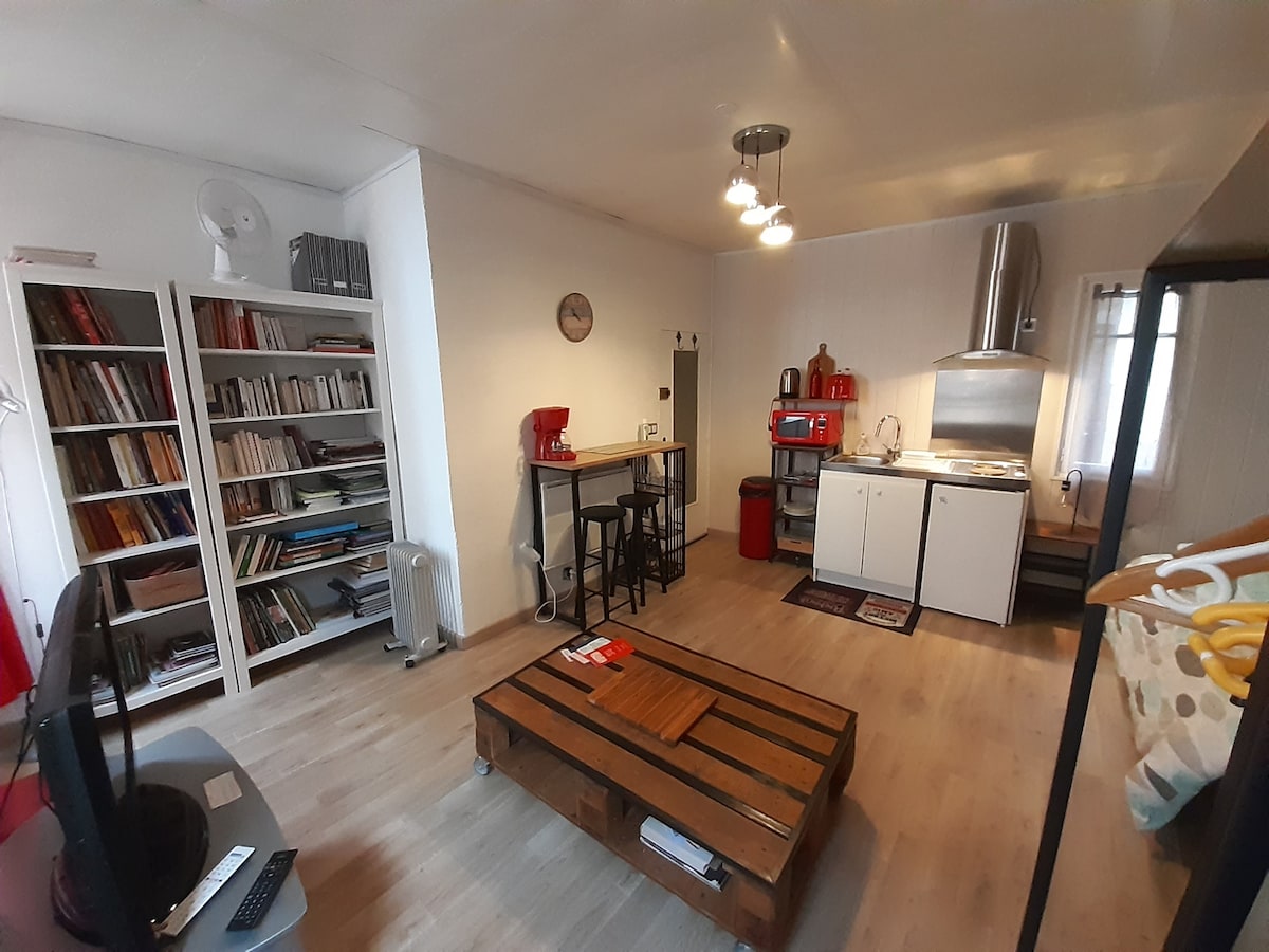 An open studio space features a dining area with high stools beside a compact kitchen. A bookshelf filled with various books is located against one wall. A wooden coffee table rests in the center, complemented by light wood flooring and bright wall accents.