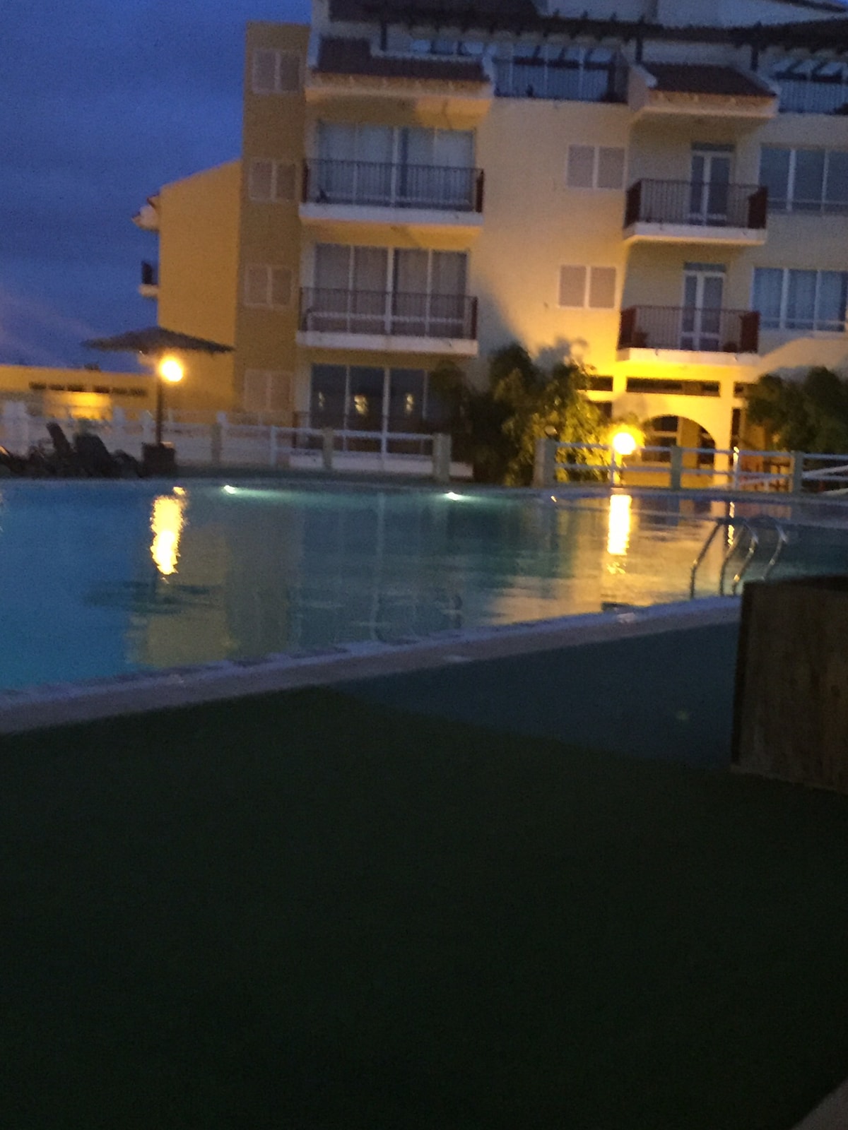 A nighttime view of the pool area reflects soft lighting from surrounding buildings and poolside lamps. The water surface shimmers under the subdued illumination, while the structure's balconies overlook the serene setting, creating a calm and inviting atmosphere.