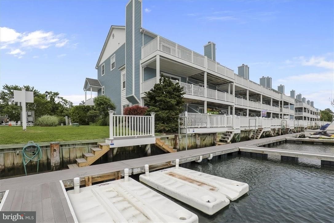 The townhouse complex features multiple levels with private decks overlooking the water. A wooden dock extends from the property, with two floating docks situated nearby. Lush greenery surrounds the area, adding to the natural setting along the waterfront.