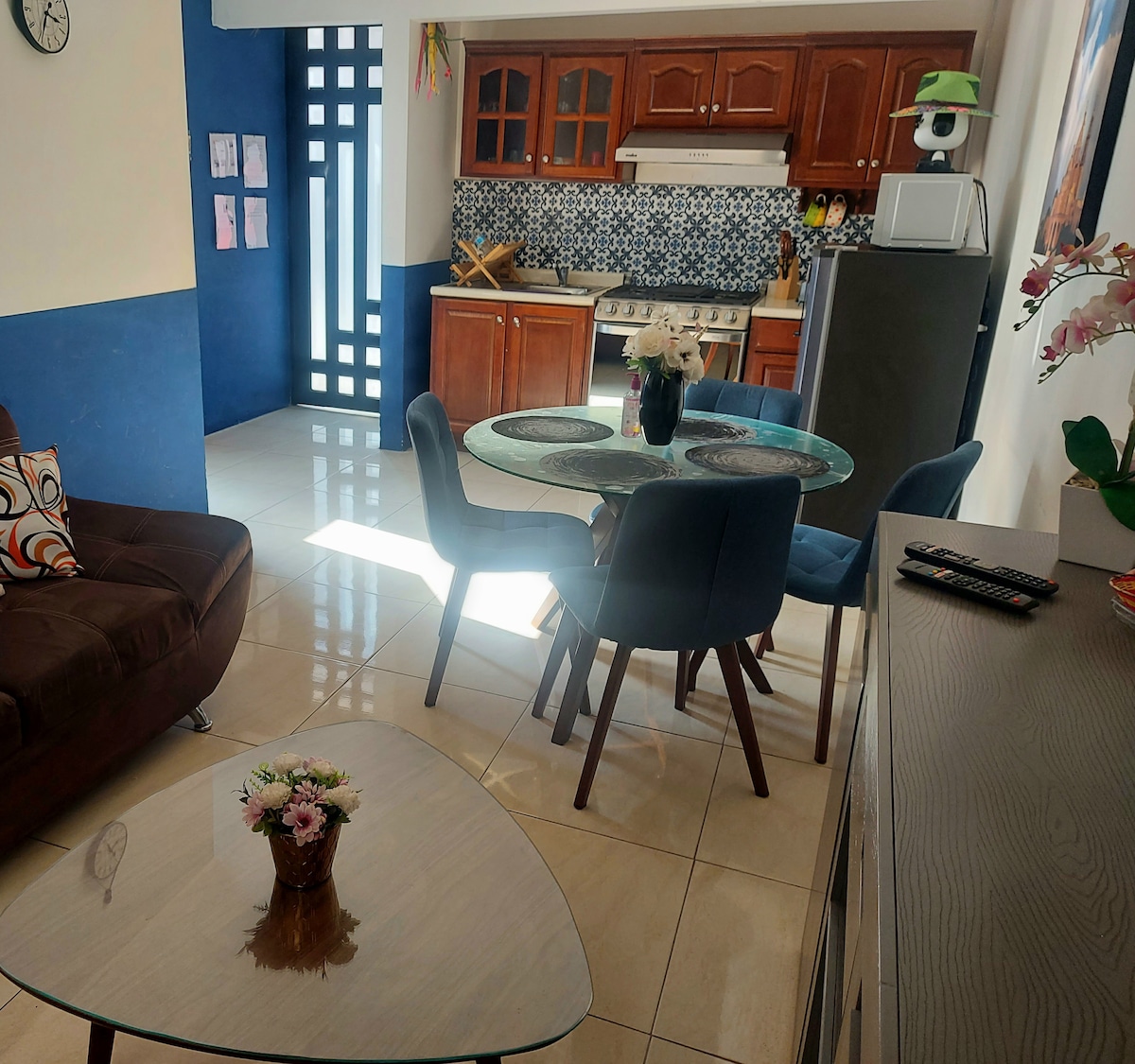 A well-lit living area features a round dining table surrounded by upholstered chairs. The kitchen showcases wooden cabinetry and colorful tile backsplash. A comfortable couch and elegant coffee table complete the space, which is complemented by sunlight streaming through the windows.