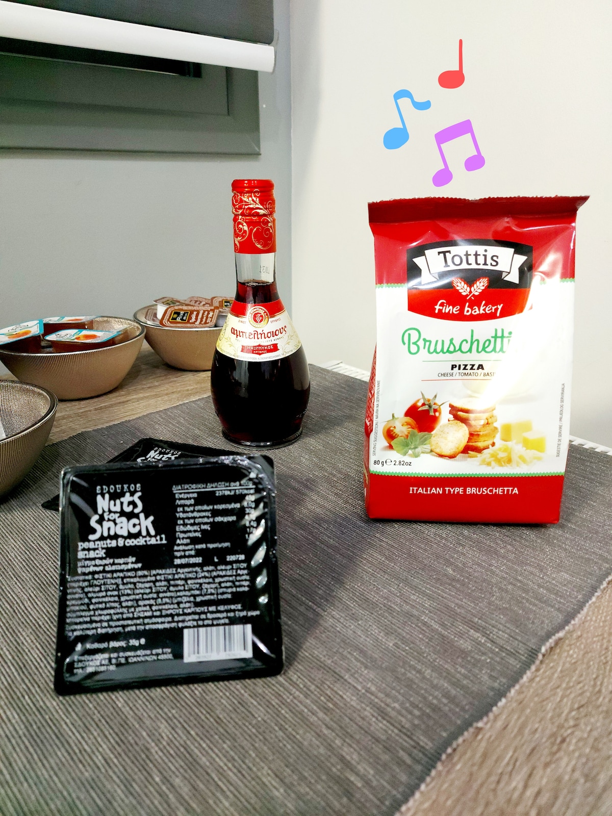 A table is set with a textured tablecloth featuring an assortment of snacks. A bottle of sauce sits alongside a package of bruschetta and a small bag of nuts, offering a variety of flavors for guests to enjoy.