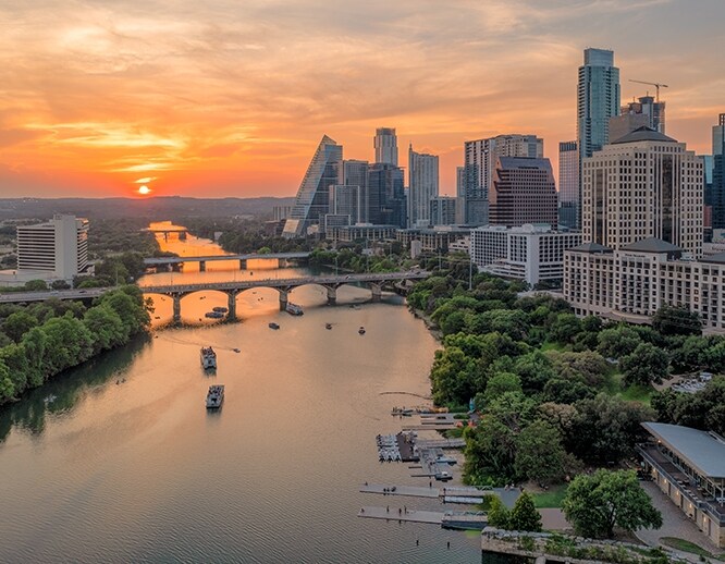 A panoramic view of Austin's skyline is captured at sunset, with vibrant orange and pink hues reflected on the water. The cityscape features tall buildings alongside the Colorado River, where boats can be seen leisurely floating near the lush green banks.