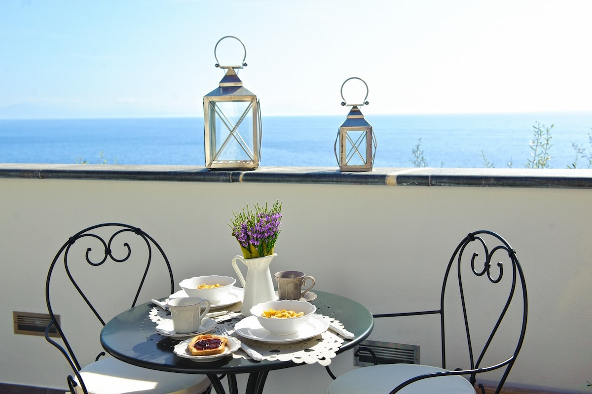 A charming outdoor terrace features a small round table set with two chairs. Fresh lavender is arranged in a white vase, accompanied by breakfast dishes and cups. Two lanterns are placed on the ledge, overlooking a serene view of the sea.