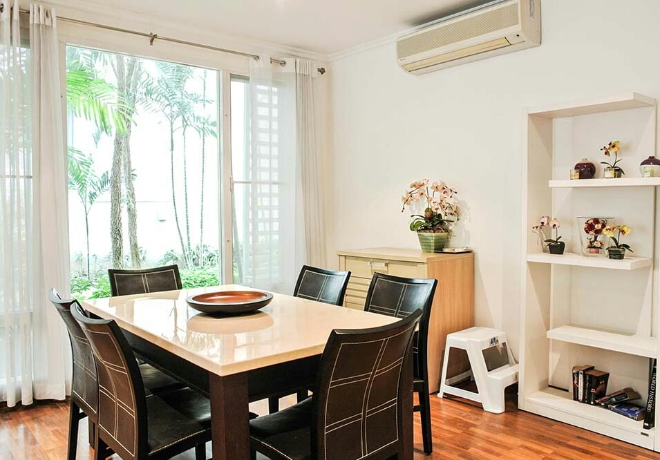 A dining area is presented with a large, square table surrounded by six black chairs. Natural light filters through a large window, illuminating the room. A stylish cabinet with decorative items stands against one wall, while a small shelf adds character with additional plants.
