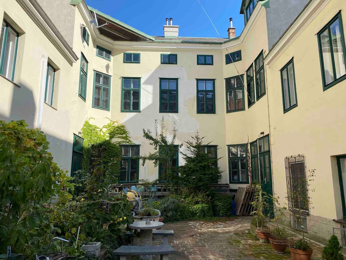 A serene courtyard area is surrounded by the exterior of a historic building. Green windows provide a view into the space, while greenery and potted plants add a natural element. Stone paths lead through the courtyard, creating a tranquil outdoor setting.