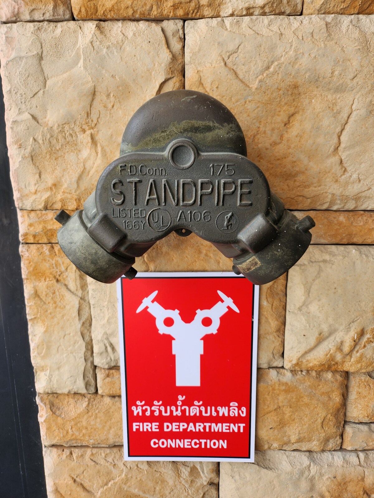 A metal standpipe connection attached to a stone wall is displayed, featuring a circular design with a marking indicating its specifications. Below it, a red sign with white graphics and text signals the fire department connection, ensuring safety and compliance.