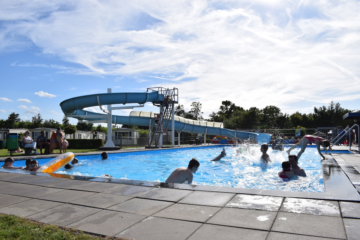 A swimming pool area features a water slide and a spacious deck, with children and families enjoying the water. Surrounding greenery enhances the outdoor atmosphere, while a clear sky and soft clouds add to the sunny ambiance. Safety measures are visible with a lifeguard tower nearby.