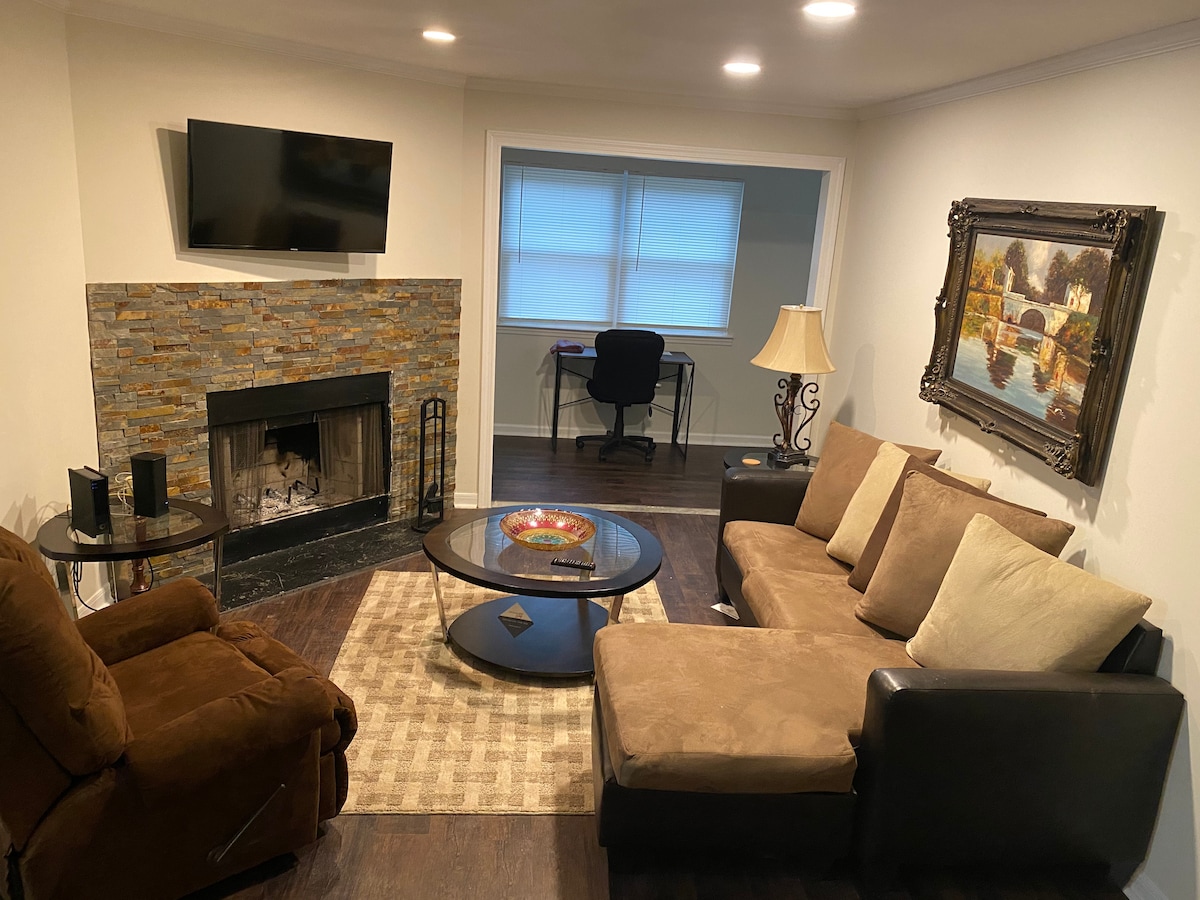 A comfortable living area features a sectional sofa and a recliner chair, with a stone fireplace as a focal point. A television is mounted above the fireplace. A small desk and chair sit against the wall, and large windows allow natural light to enter.