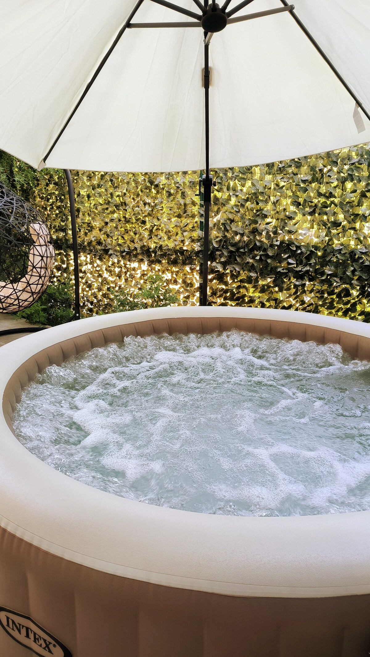 A private hot tub sits under a large umbrella, surrounded by lush greenery. Bubbles create a soothing effect on the water surface, while a soft light filters through the leaves, enhancing the serene atmosphere of the outdoor space.