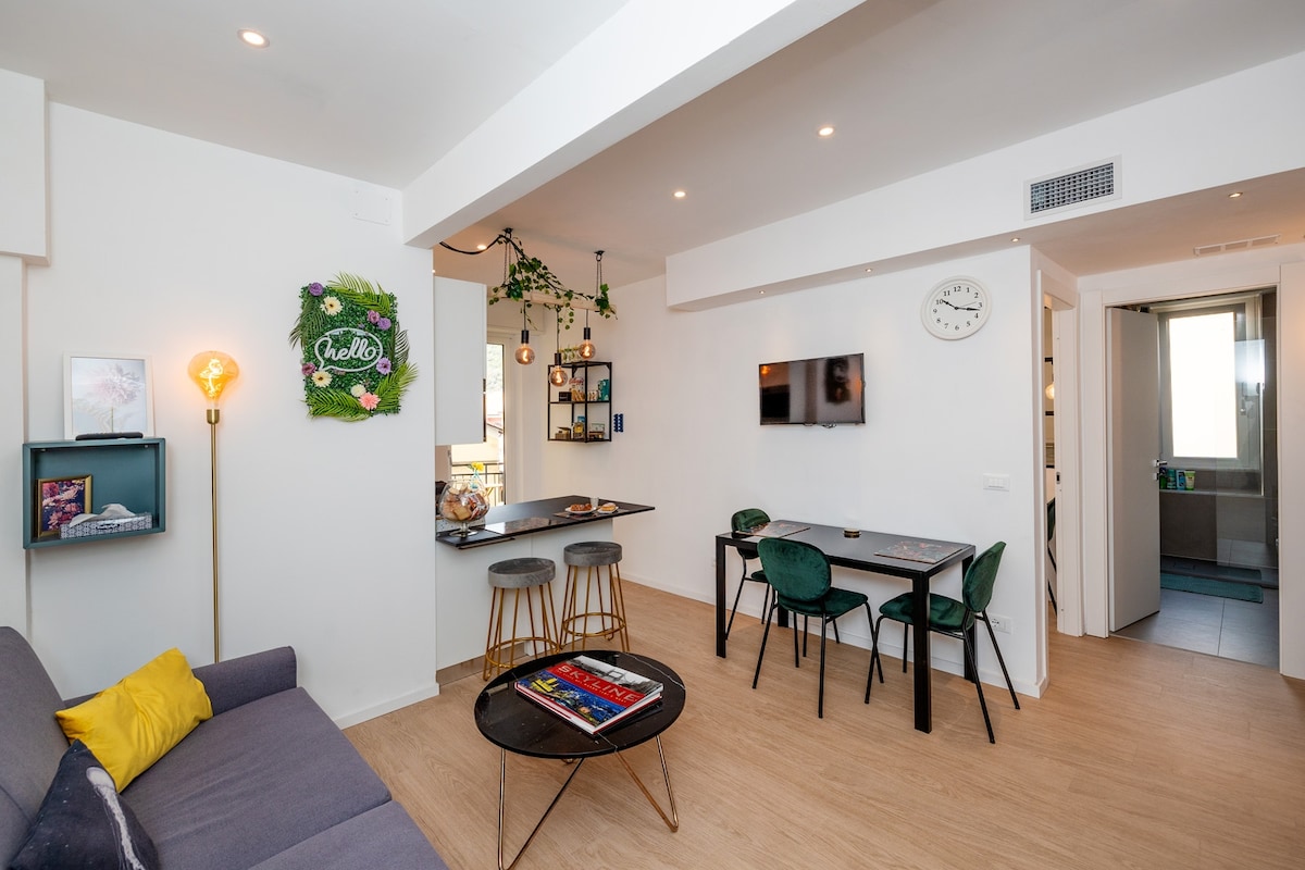 An open-concept living area features a cozy seating arrangement with a sofa and vibrant cushion. A round table with chairs sits nearby, while a small bar with stools is positioned next to a wall adorned with decorative plants. Natural light enters through the adjacent room.