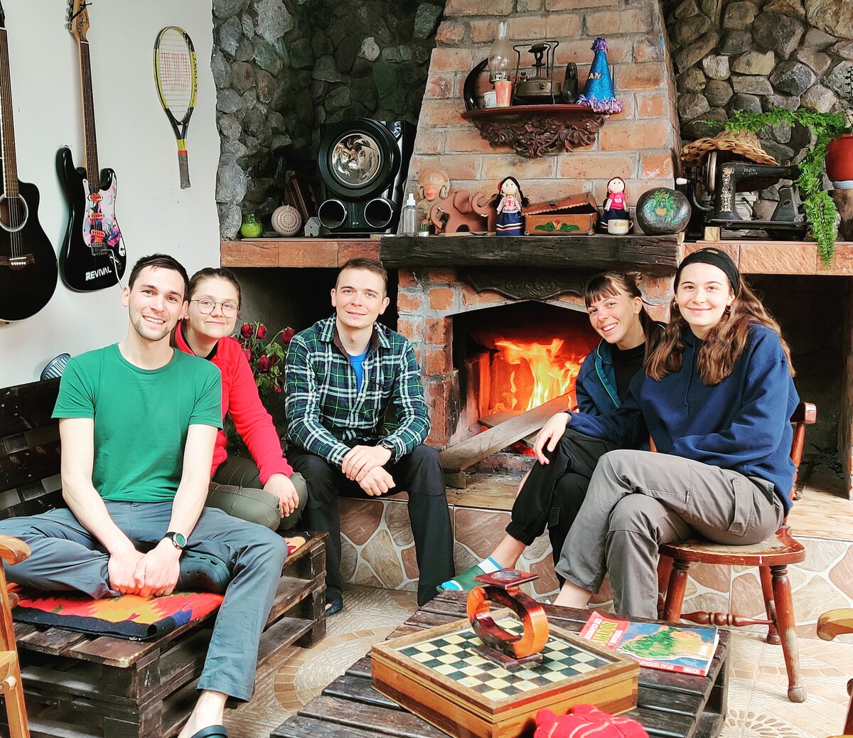 A cozy seating area features a stone fireplace with a warm glow. Several individuals sit on rustic wooden chairs around a checkered board game, surrounded by local decor items. Guitars are displayed on the wall, enhancing the welcoming space.
