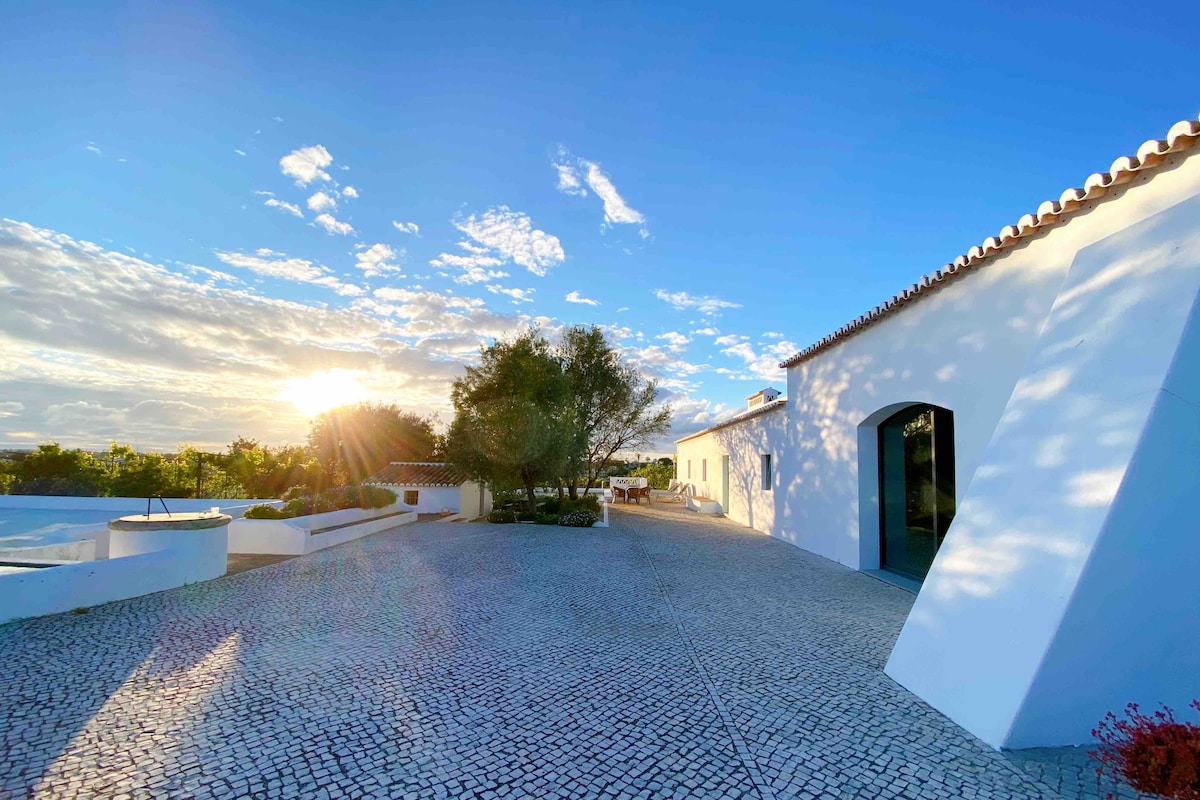 A spacious outdoor area showcases a cobblestone pathway leading towards a white building. Lush greenery borders the space, and gentle slopes create a natural ambiance. The warm glow of the setting sun casts a serene light over the surroundings, enhancing the tranquil atmosphere.