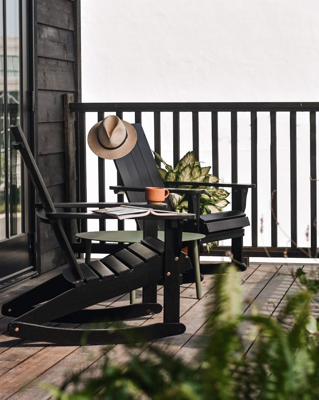 A semi-private porch is shown with two black rocking chairs positioned near a small table. A wide-brimmed hat rests atop one chair, and a potted plant adds a touch of greenery. The light-colored wall in the background complements the natural materials.