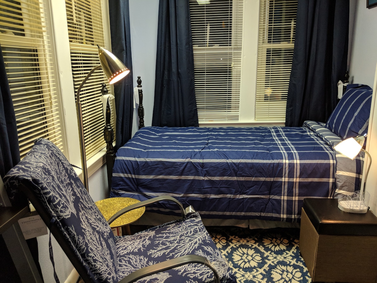 A private room features a compact bed dressed in blue and white striped linens, alongside a decorative armchair with intricate patterns. Tall windows allow natural light to brighten the space, while dark curtains frame the room, creating a cozy ambiance.