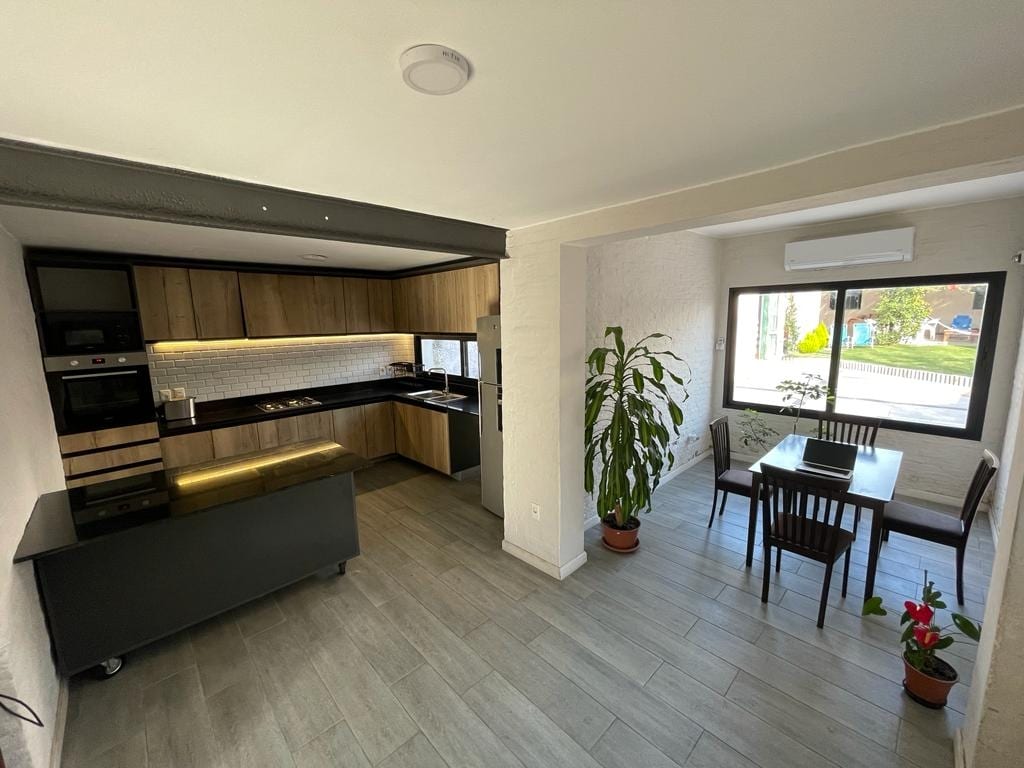 A modern kitchen and dining area are shown, featuring sleek cabinetry and appliances. A small dining table with four chairs is positioned near a large window, allowing natural light to fill the space. Indoor plants add a touch of greenery to the atmosphere.