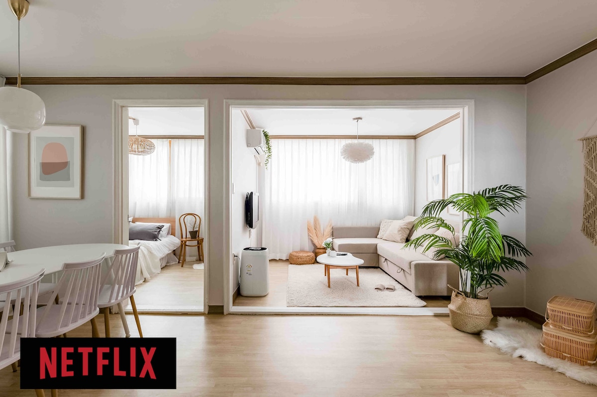 A spacious living area includes a comfortable sofa and coffee table, with natural light filtering through sheer curtains. The adjacent dining area features a light-colored table with accompanying chairs. Green plants add a touch of nature, enhancing the overall ambiance.