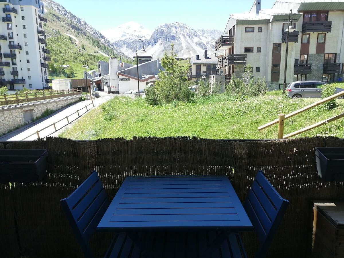 A simple terrace area features a blue wooden table accompanied by two matching chairs. In the background, a grassy space and mountain scenery are visible, with buildings framed against a clear sky.