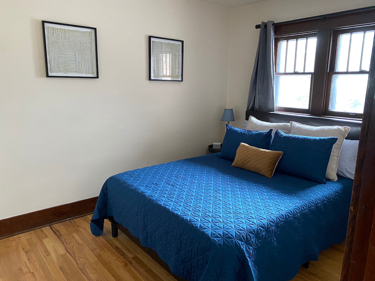 A bedroom features warm wood flooring and large windows that allow natural light to enter. A spacious bed is dressed in a textured blue quilt, complemented by decorative pillows. Two framed artworks adorn the walls, adding a touch of character.