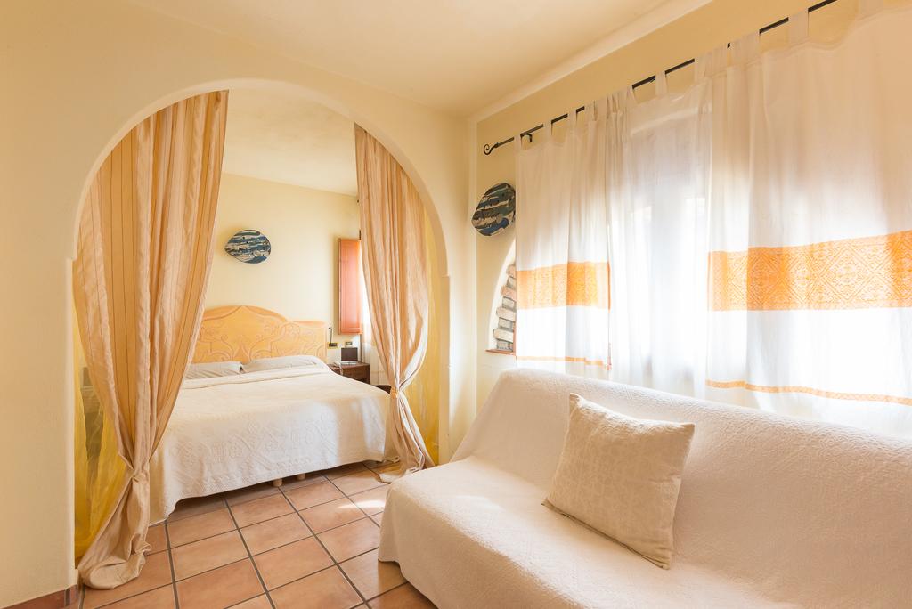 A cozy room features an arched entrance leading to a spacious bed adorned with a light-colored coverlet. Soft curtains with orange accents frame the window, allowing natural light to brighten the area. A simple white sofa with a cushion is positioned nearby.
