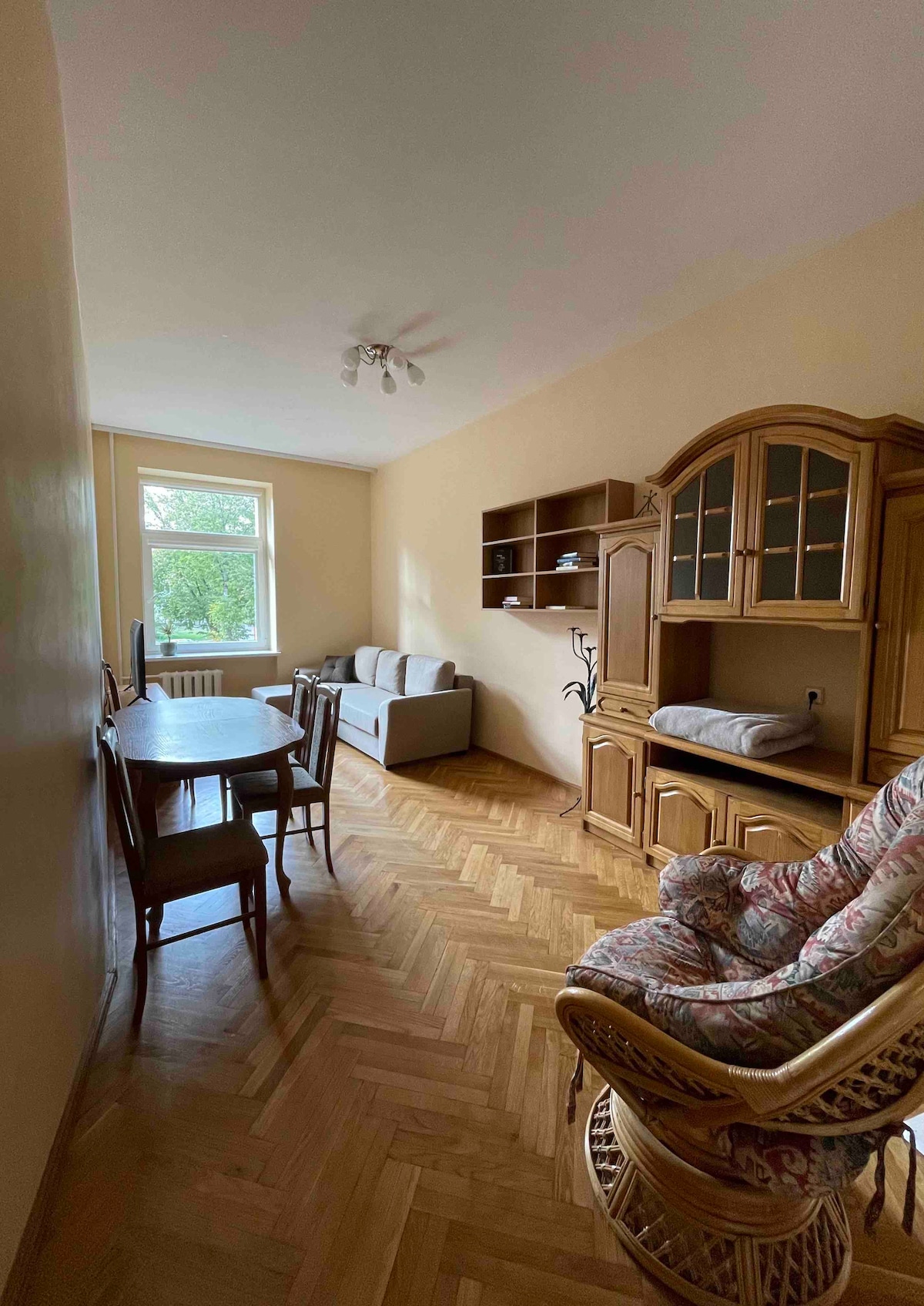 A warmly decorated living space features a grey sofa and a wooden dining table surrounded by four chairs. Light-colored walls and hardwood flooring create a bright atmosphere. A stylish cabinet displays decorative items, and a chair with natural fibers adds a cozy touch.