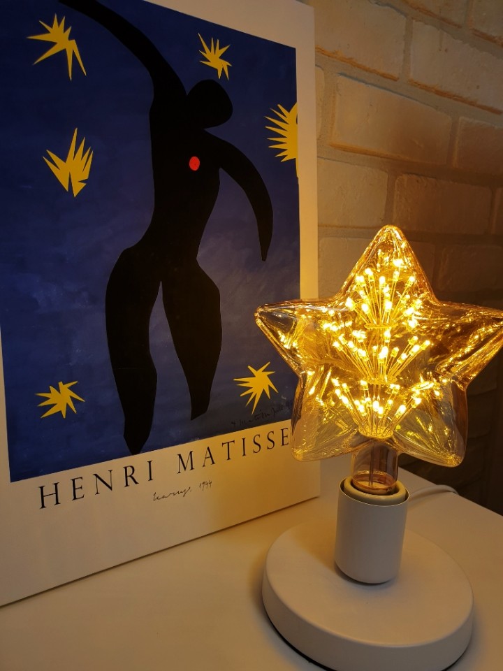 A star-shaped lamp emits a warm glow, positioned next to a colorful art print featuring the silhouette of a figure by Henri Matisse. The print is framed by bright blue and yellow elements, adding a creative touch to the setting.