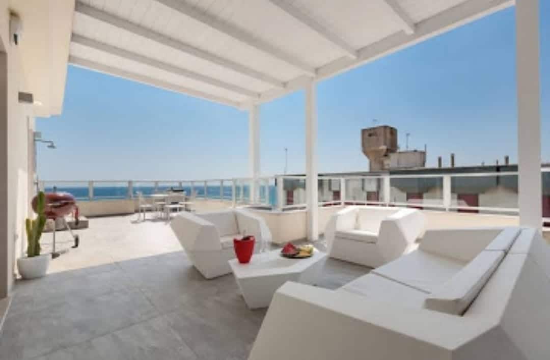 A spacious outdoor terrace is depicted, featuring modern white furniture arranged for relaxation. A table with chairs is visible, alongside a grill. The backdrop includes a clear blue sky and the view of the sea, enhancing the outdoor experience.