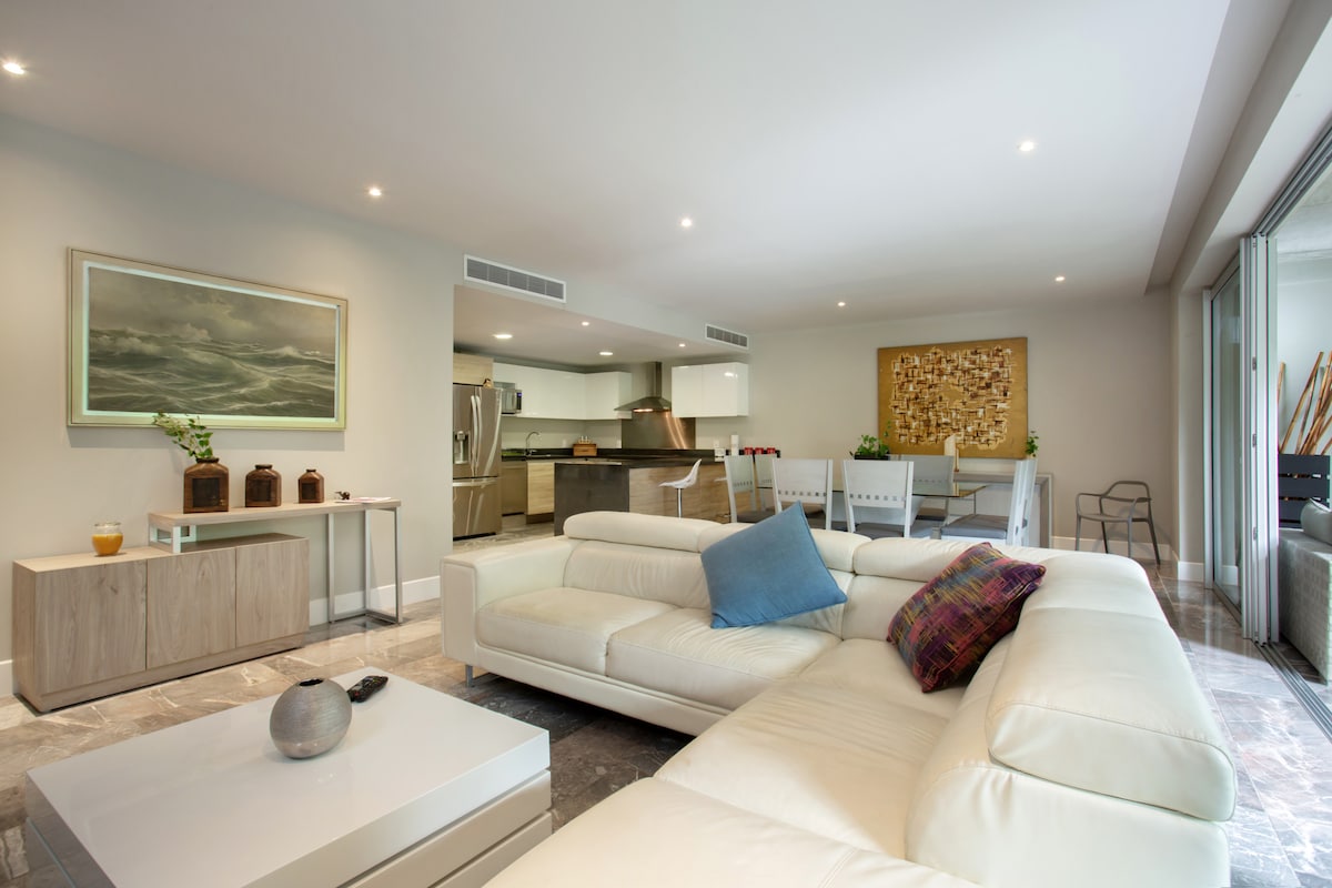 A spacious living area is highlighted by a sectional white sofa adorned with a colorful accent pillow. A neutral-toned art piece and a coffee table are positioned in front of the sofa, while a modern kitchen and dining area are visible in the background.