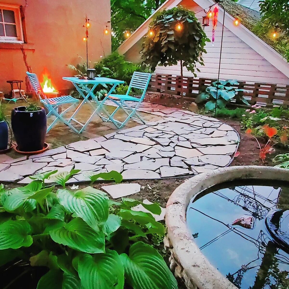 A charming outdoor patio area features a stone pathway, small table and chairs in blue, and a serene pond with visible reflections. Soft lighting from string lights and lanterns enhances the outdoor space, surrounded by lush greenery and garden elements.