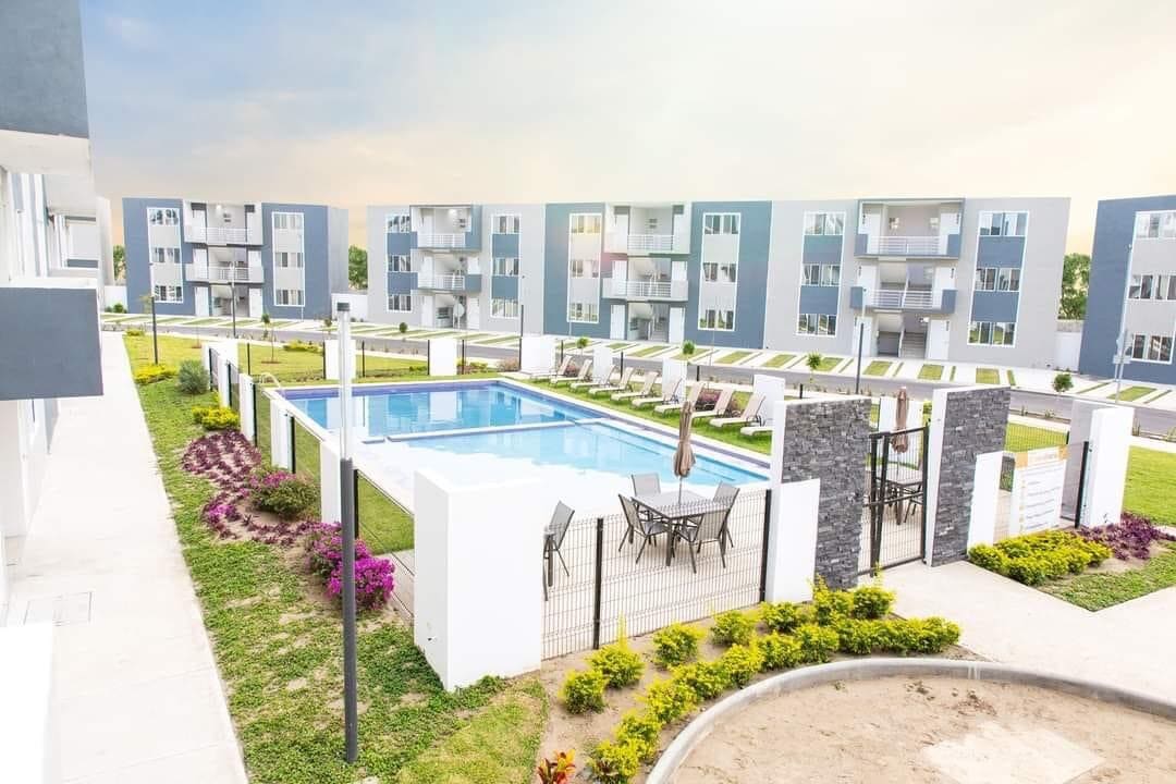 A large swimming pool is surrounded by a well-maintained green area, featuring lounge chairs and outdoor tables. Modern residential buildings are visible in the background, complemented by neatly arranged flowering plants along the pathways.