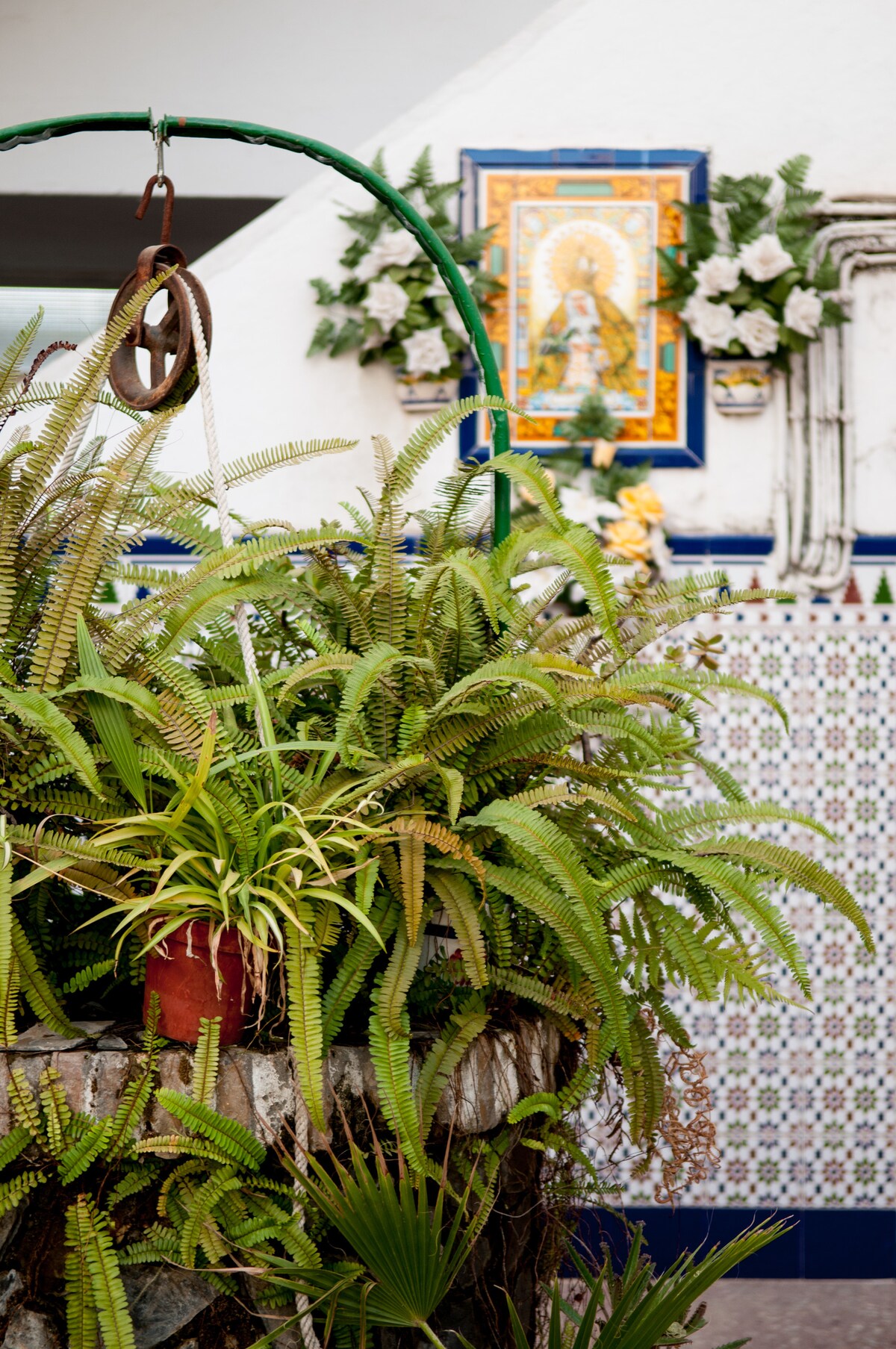 A vibrant display of greenery is featured, with lush ferns and potted plants arranged around a stone structure. In the background, a colorful mural is visible, surrounded by intricate tile work and floral decor, creating an inviting atmosphere that reflects local craftsmanship.