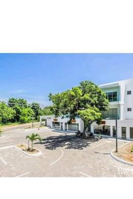 The exterior showcases a modern building surrounded by landscaped greenery and a spacious parking lot. A large tree provides shade near the entrance, while clear blue skies create a bright and open atmosphere.