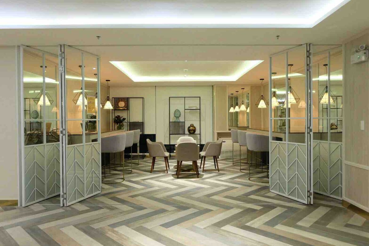 The lounge area features a modern design with patterned herringbone flooring and soft, neutral tones. Elegant lighting fixtures hang above circular seating arrangements, complemented by decorative shelving. Two wide doors with geometric patterns provide access to the space, enhancing the open feel.