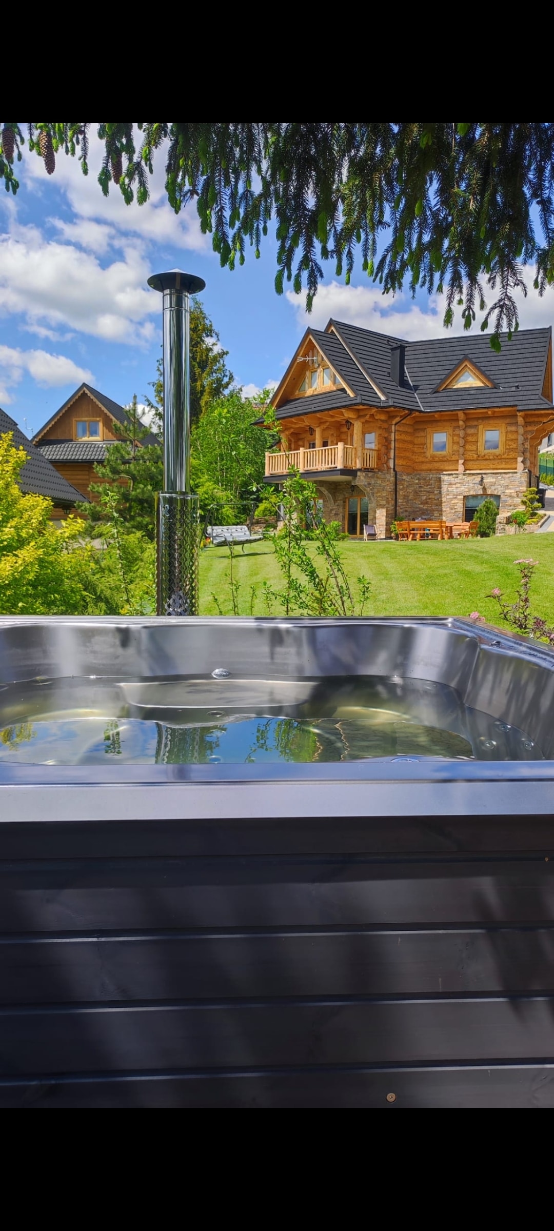 A hot tub sits in the foreground, surrounded by lush greenery. In the background, a wooden house with a sloped roof is visible amidst a vibrant landscape, with clear skies above adding to the serene ambiance.