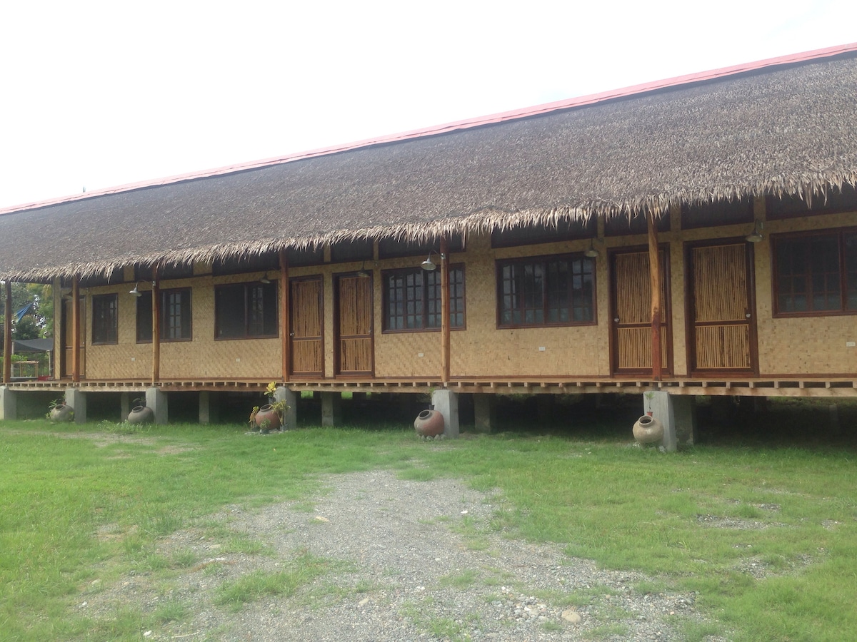 A row of modern bamboo hotel units is presented, featuring thatched roofs and wooden doors. The units are elevated on stilts, surrounded by grassy areas. Each unit has a small patio, and decorative clay pots are positioned at the base of the stairs.