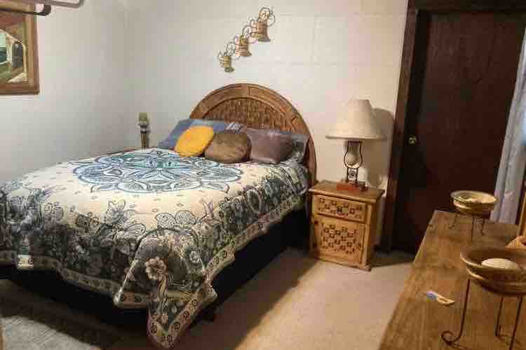 A cozy bedroom features a spacious bed dressed in a patterned quilt with decorative pillows. A bedside table with a lamp is positioned next to the bed. A wooden dresser with bowls is visible, and artwork hangs on the walls, contributing to the room's character.