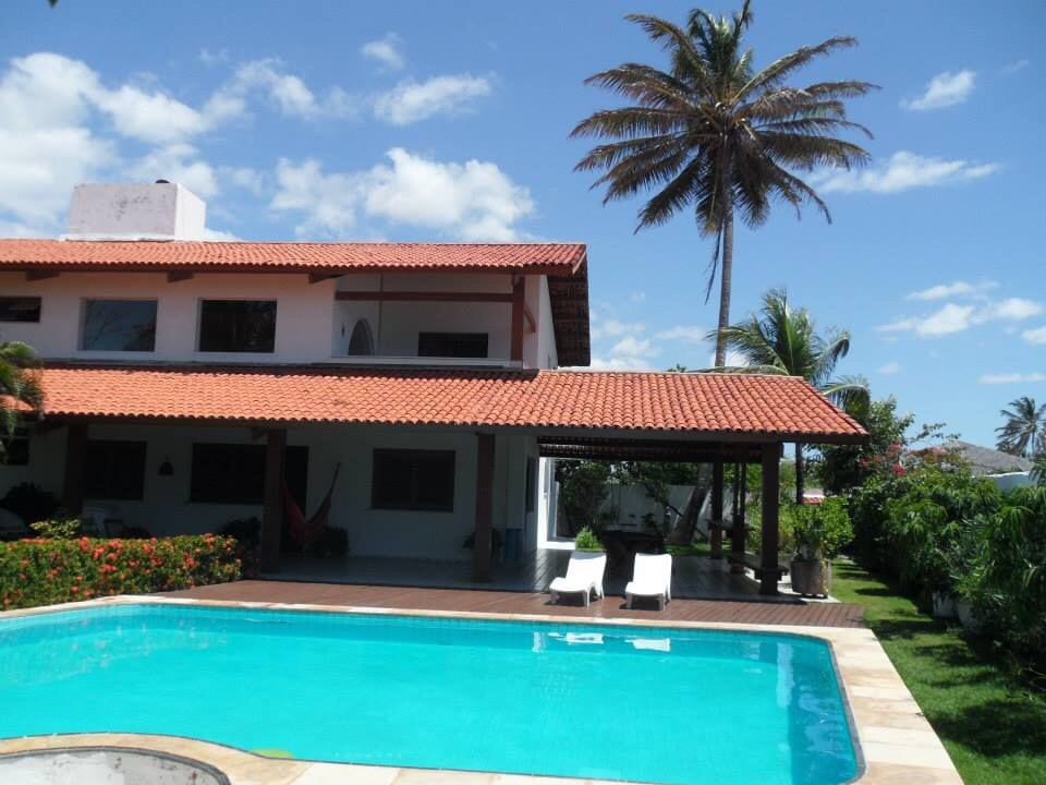 A newly renovated villa is presented, featuring a spacious pool surrounded by a wooden deck. Lush greenery is visible in the garden, and a shaded area with lounge chairs can be seen. The structure offers a warm exterior with a terracotta roof.