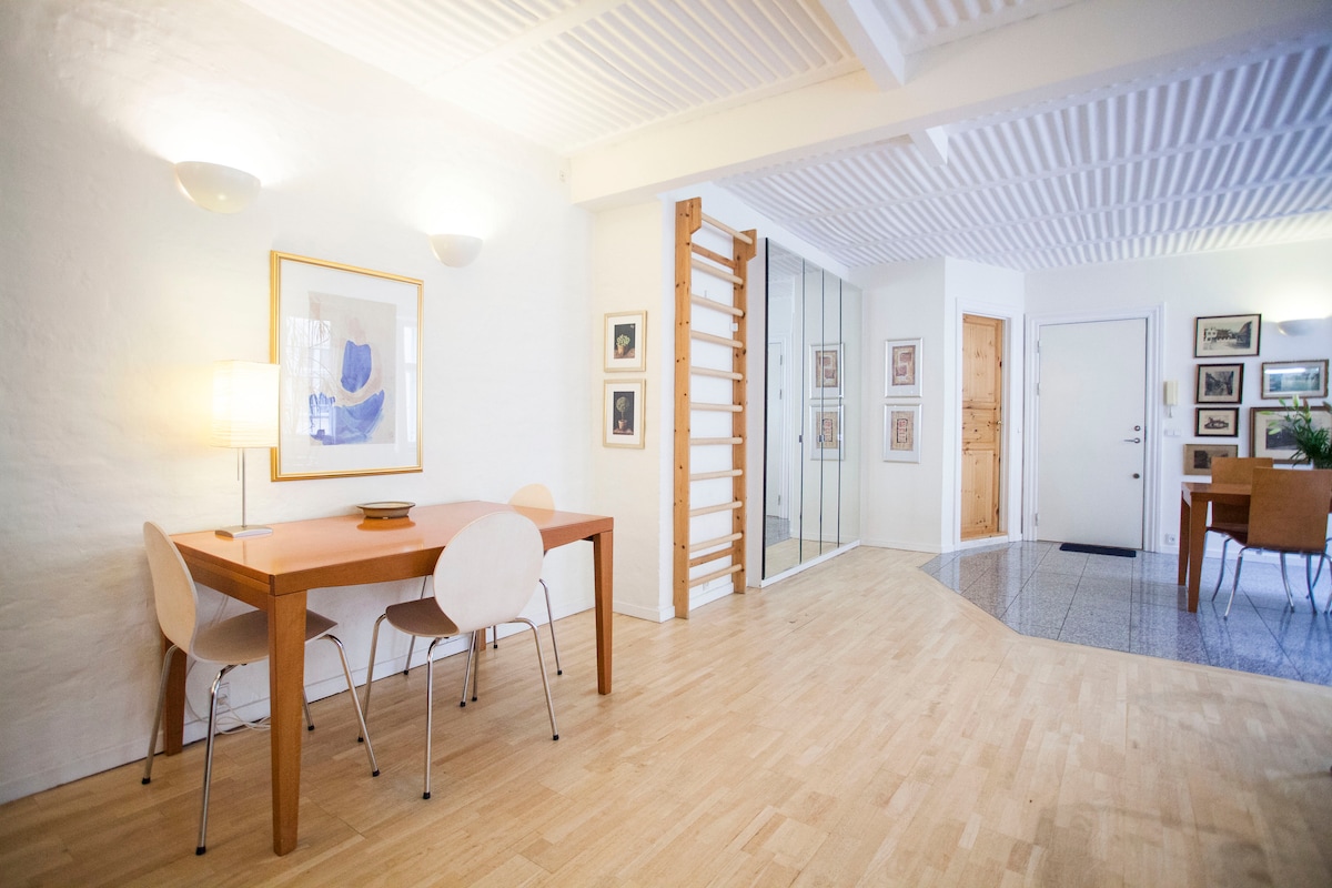 An open dining area featuring a wooden table surrounded by white chairs is illuminated by soft lighting. The space extends into a light-filled hallway with framed artwork on the walls and a staircase leading to an upper level, showcasing a blend of modern and classic design.
