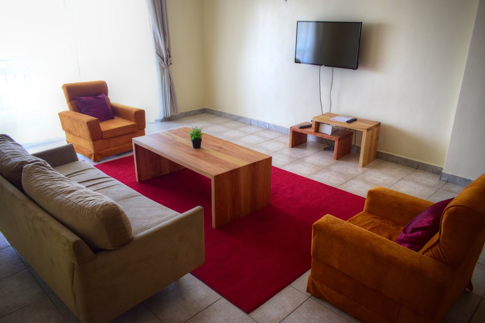 A furnished living area is presented with a beige sofa and two orange armchairs, arranged around a wooden coffee table on a red area rug. A mounted television is positioned on the wall opposite the seating, with light entering through sheer curtains.