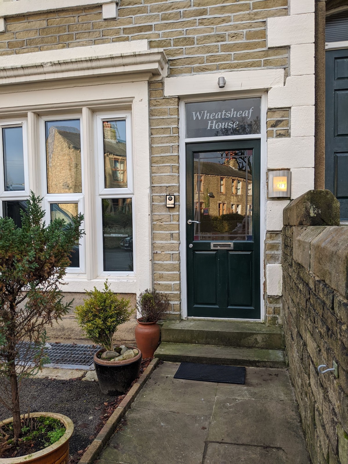 The entrance to Wheatsheaf House is marked by a dark green door, framed by light-colored stone walls. A small garden area with potted plants adds a touch of greenery, while large windows flanking the entrance bring in natural light.