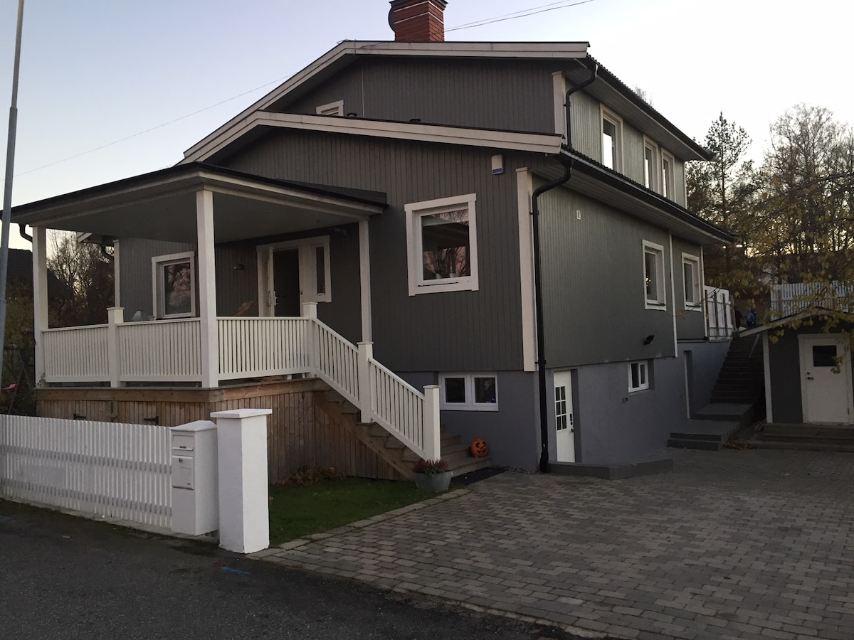 The exterior of a two-story family house is shown, featuring a gray facade with white trim. A covered porch with steps leads to the entrance, and a spacious driveway made of paving stones provides access to the property. Several windows allow for natural light.
